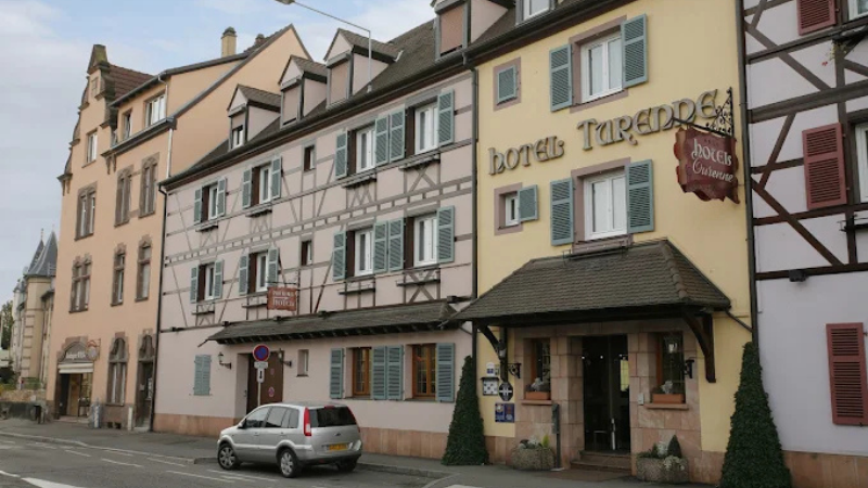 Fasad av Hotel Turenne i Colmar med traditionell korsvirkesarkitektur, pastellfärger och skylt vid entrén på en stadsgata.